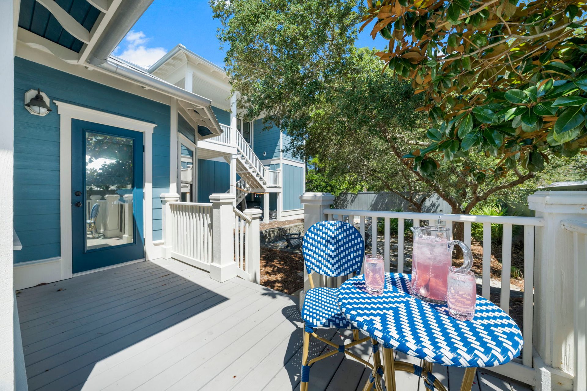 Blue porch with white railing, striped tablecloth, and pink drinks under a leafy tree