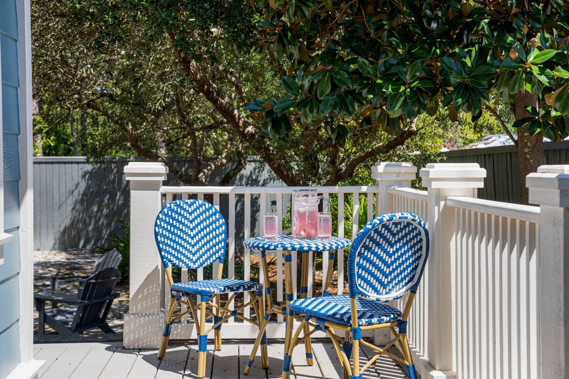 Blue wicker patio chairs and table on a sunny deck with white railings and trees beyond