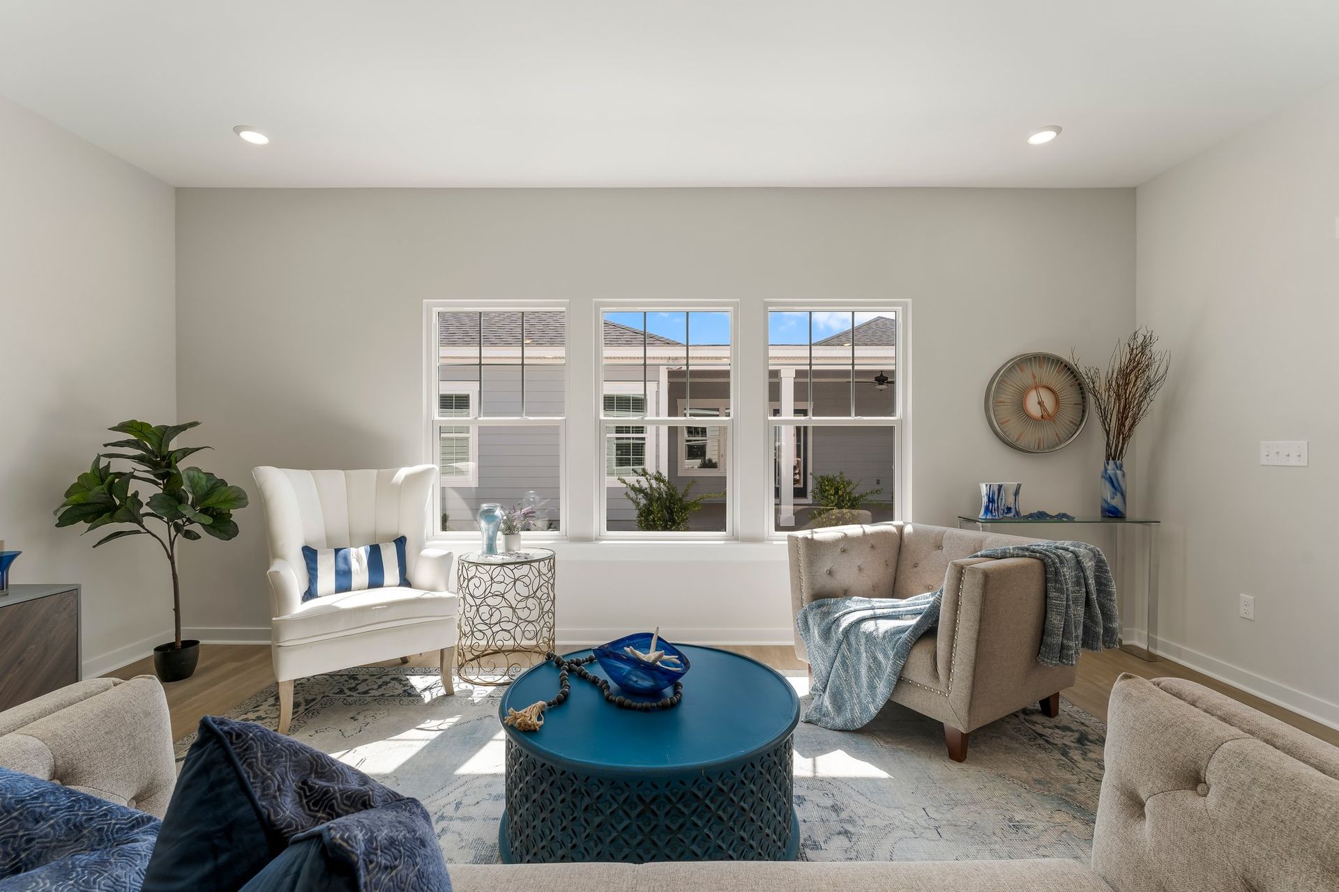 Bright living room with white chairs, blue accents, a round coffee table, and windows overlooking a patio.