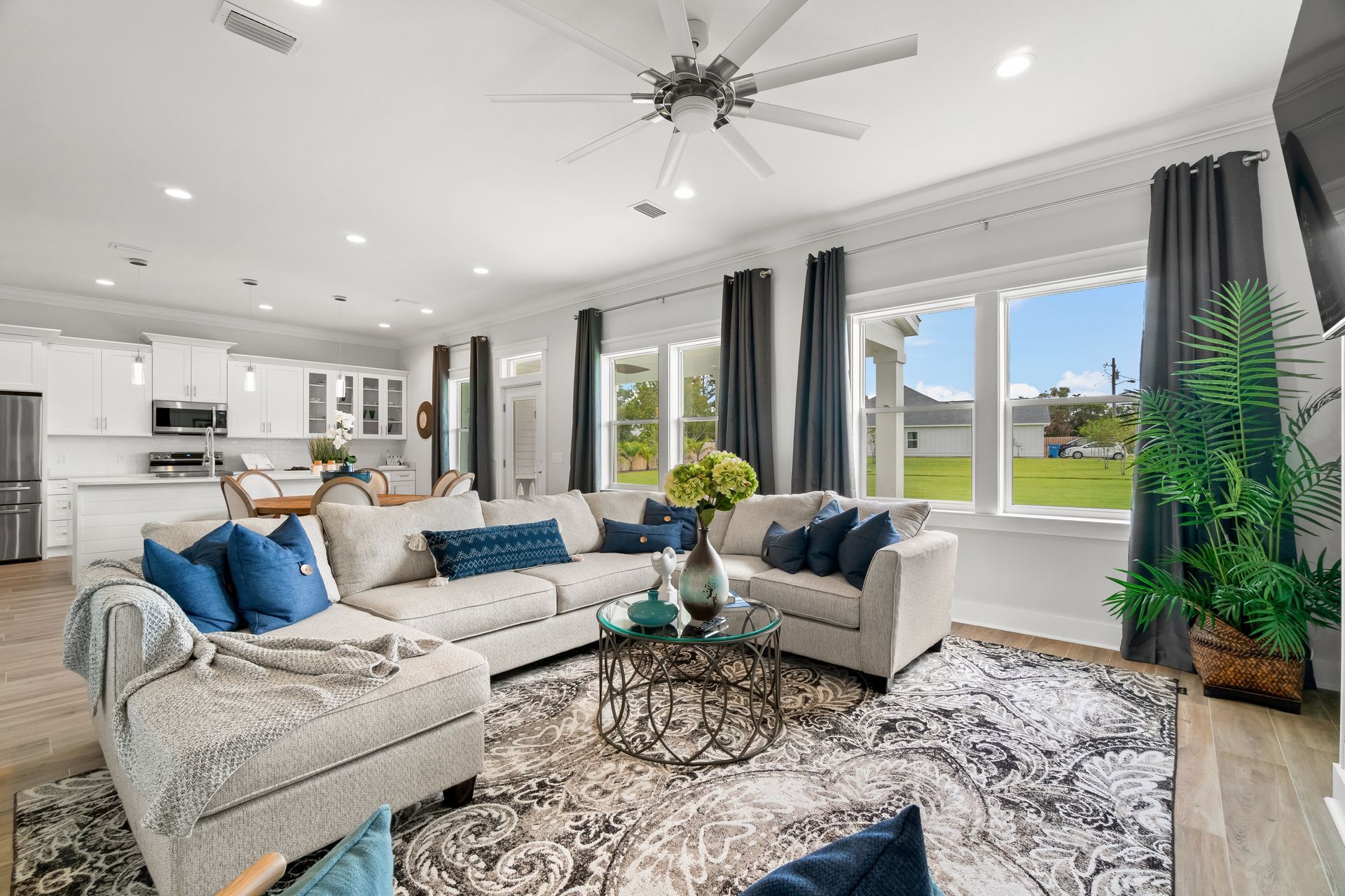 Bright modern living room with gray sectional, blue accents, large windows, and a kitchen in the background