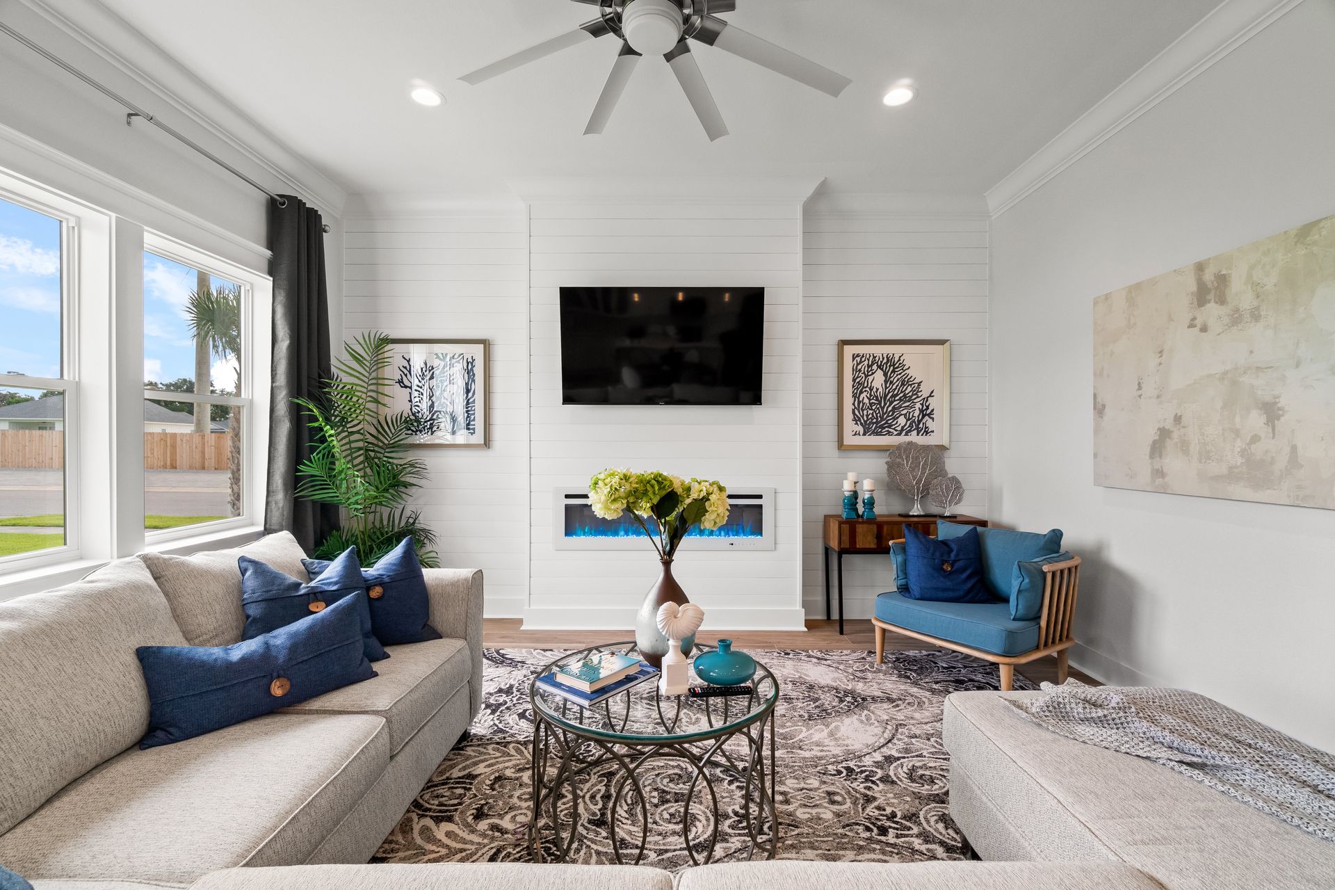 Bright living room with white shiplap walls, mounted TV, blue chairs, gray sofa, and patterned rug.