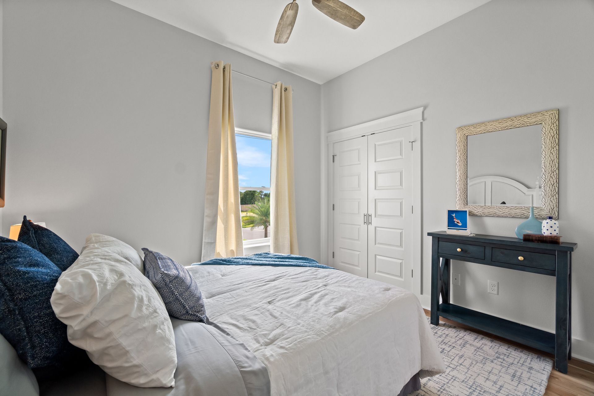 Bright bedroom with a made bed, gray walls, white door, window, and black console table with mirror.