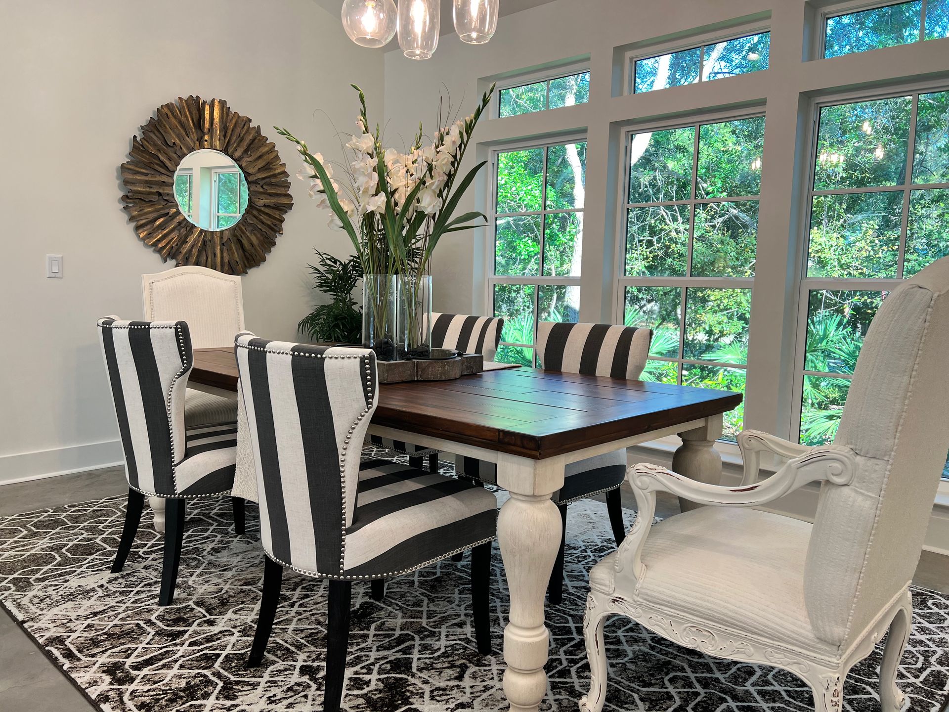 Dining room with striped chairs, dark wood table, round mirror, chandelier, and large windows overlooking greenery