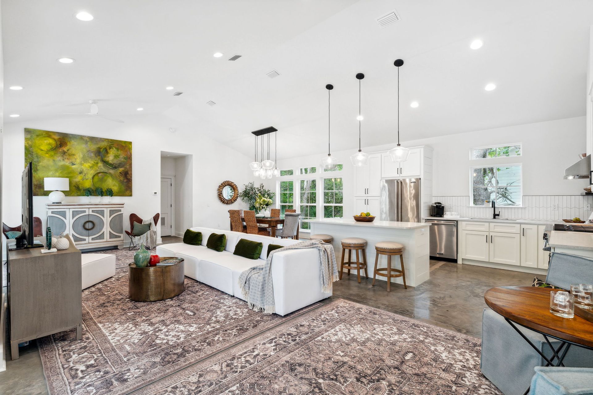 Bright open-concept living room and kitchen with white walls, hardwood floors, patterned rug, and colorful art.