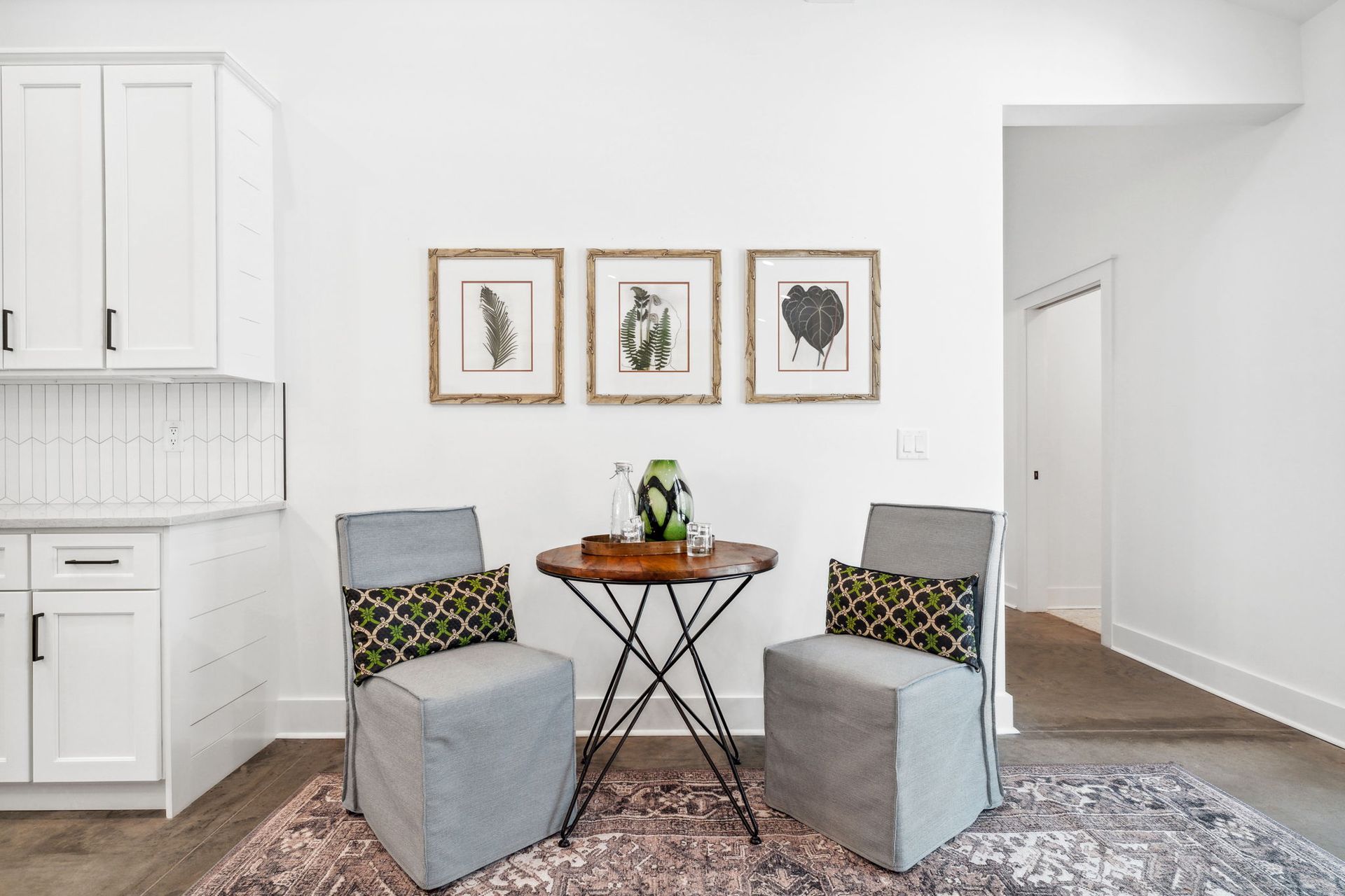 Modern living room seating area with two gray chairs, a small table, wall art, and kitchen cabinets nearby