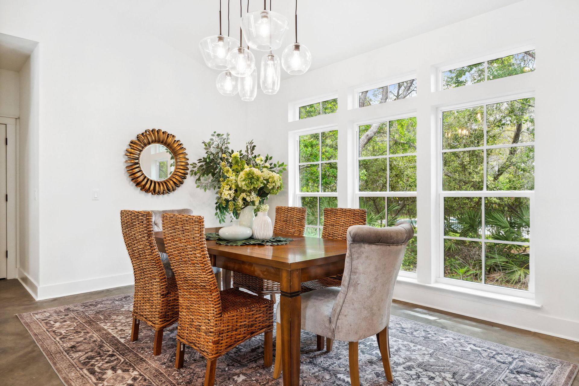 Bright dining room with wooden table, patterned chairs, chandelier, and large windows overlooking greenery
