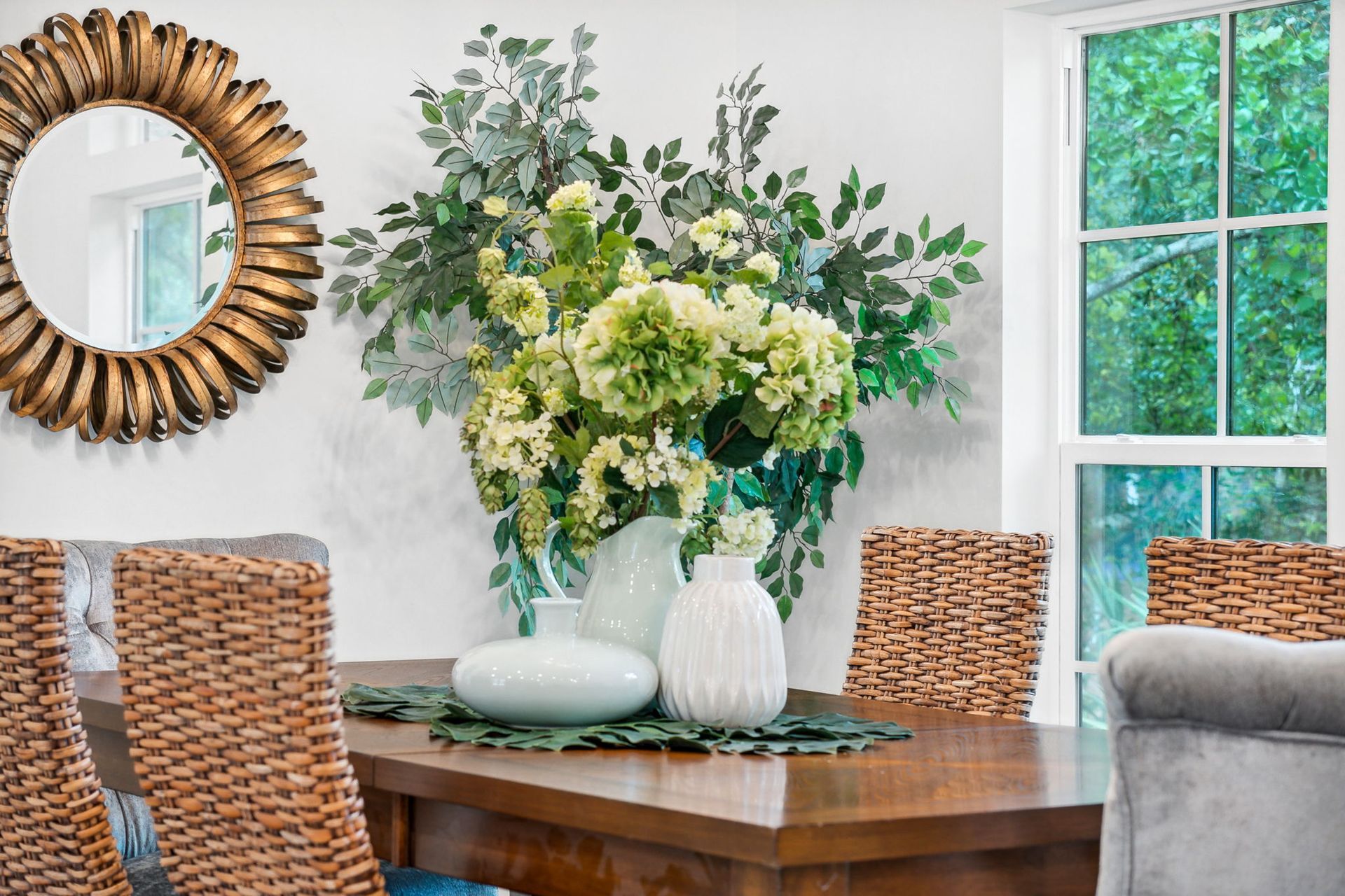 Dining table with wicker chairs, white vases, and a large green floral arrangement in a bright room
