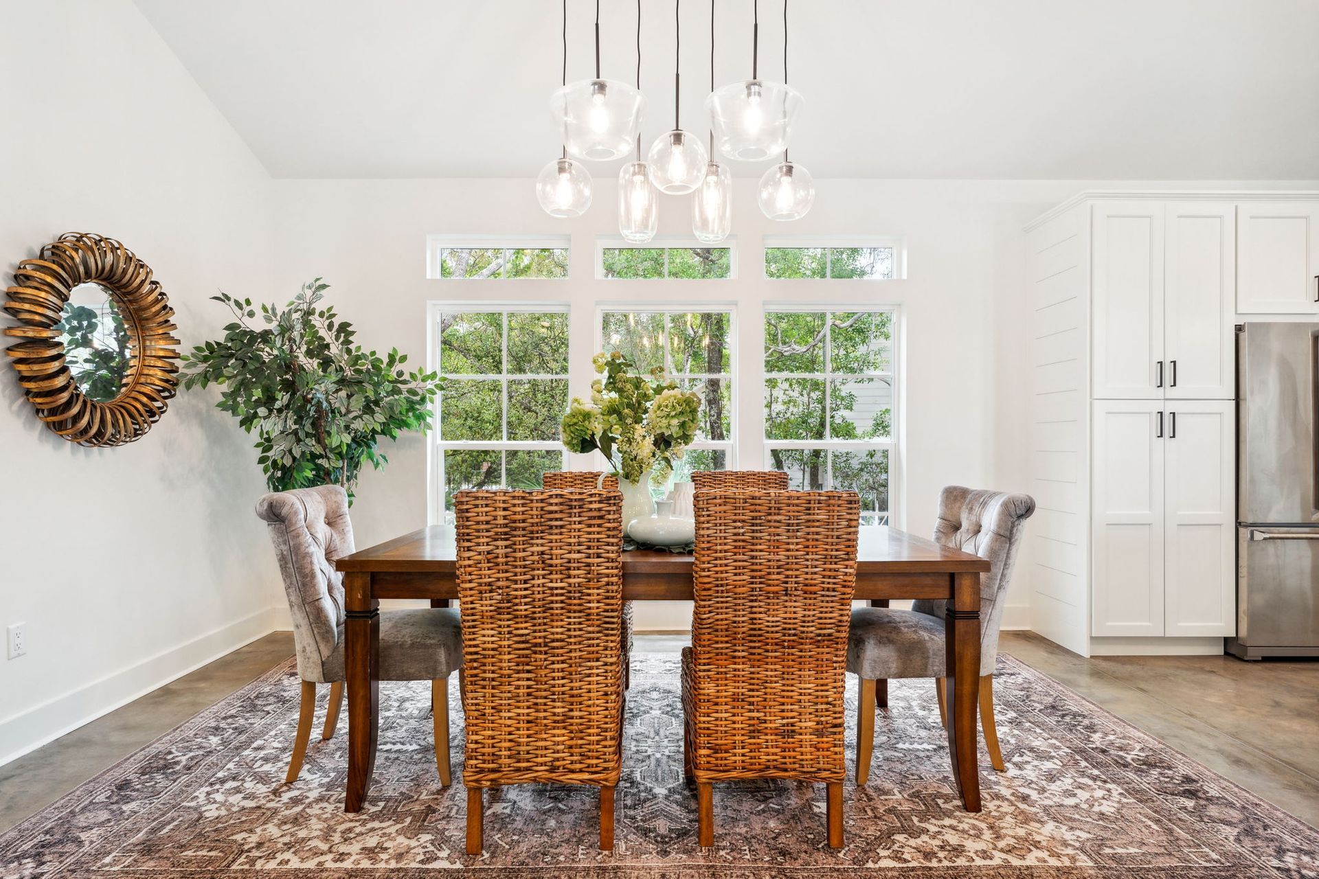 Dining room with woven chairs, wooden table, chandelier, and large windows overlooking greenery.
