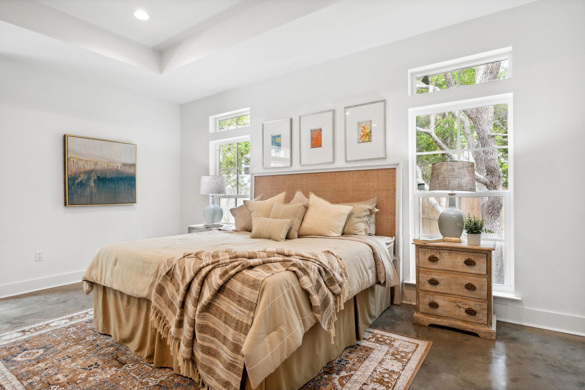 Bright bedroom with a tan bed, patterned rug, wooden nightstand, and large windows letting in natural light
