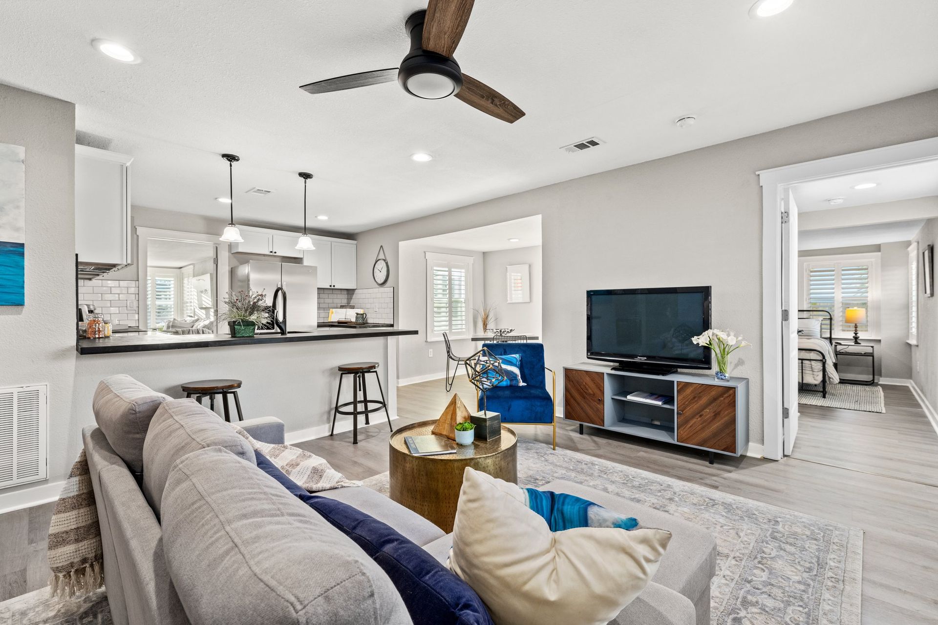 Bright open living room with gray sofa, TV, ceiling fan, and view into a modern kitchen and dining area