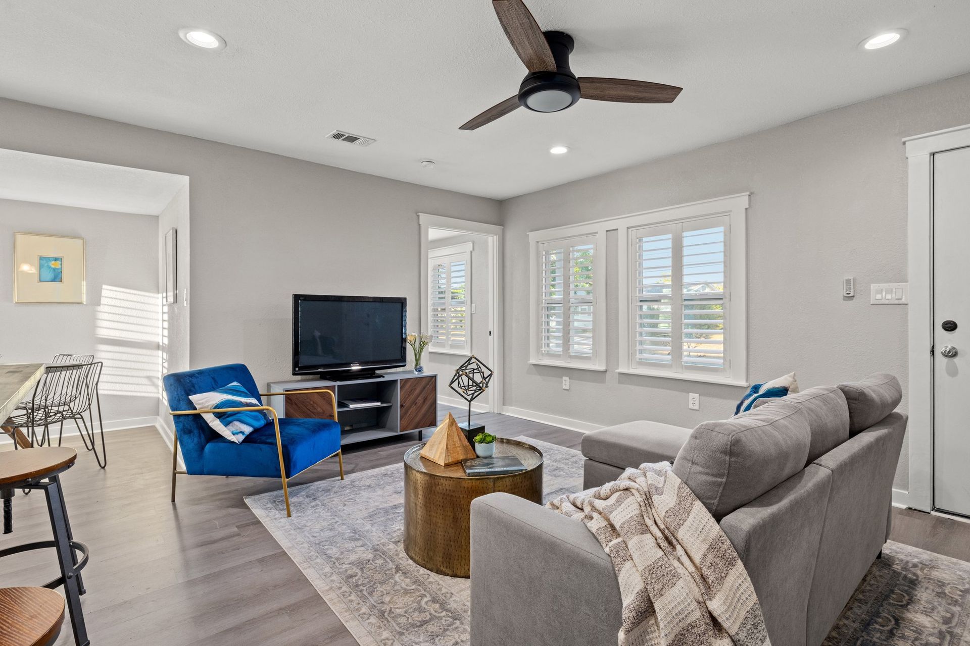 Bright living room with gray sofa, blue chairs, TV, and large windows under a ceiling fan.