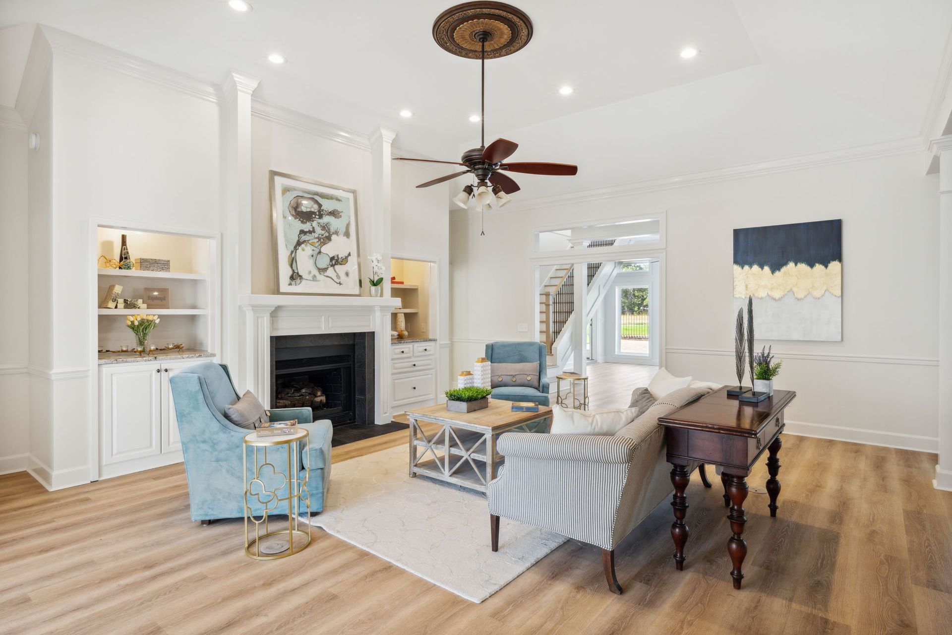 Bright living room with fireplace, blue chairs, striped ottoman, wood floors, and ceiling fan