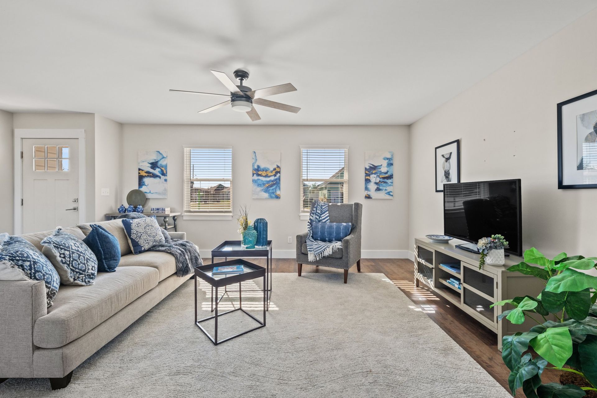Bright living room with gray sofa, blue accents, TV, ceiling fan, and large windows