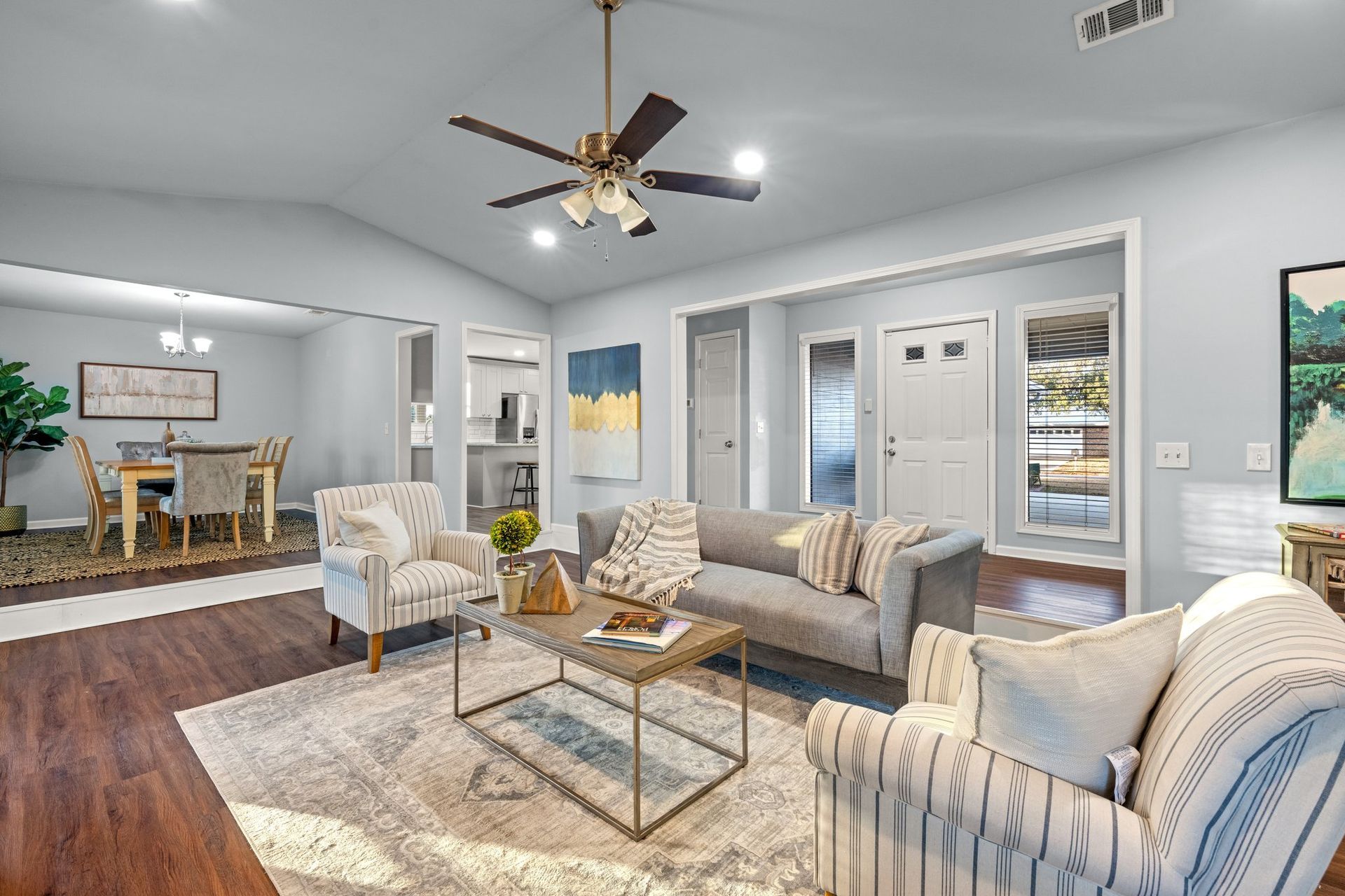 Bright living room with gray sofas, striped chairs, glass coffee table, and dining area beyond.