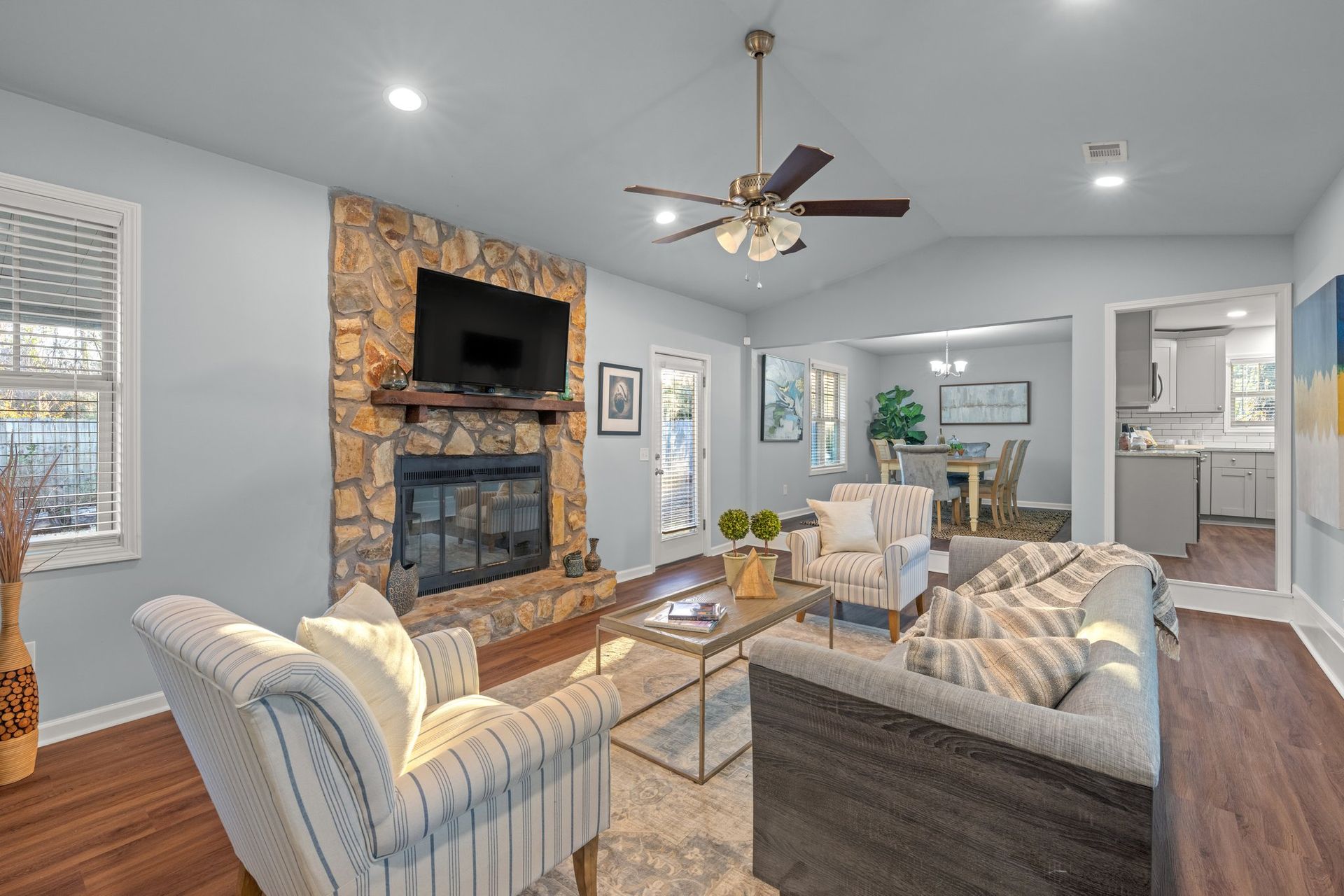 Bright living room with stone fireplace, wall-mounted TV, gray sofas, and dining area in the background