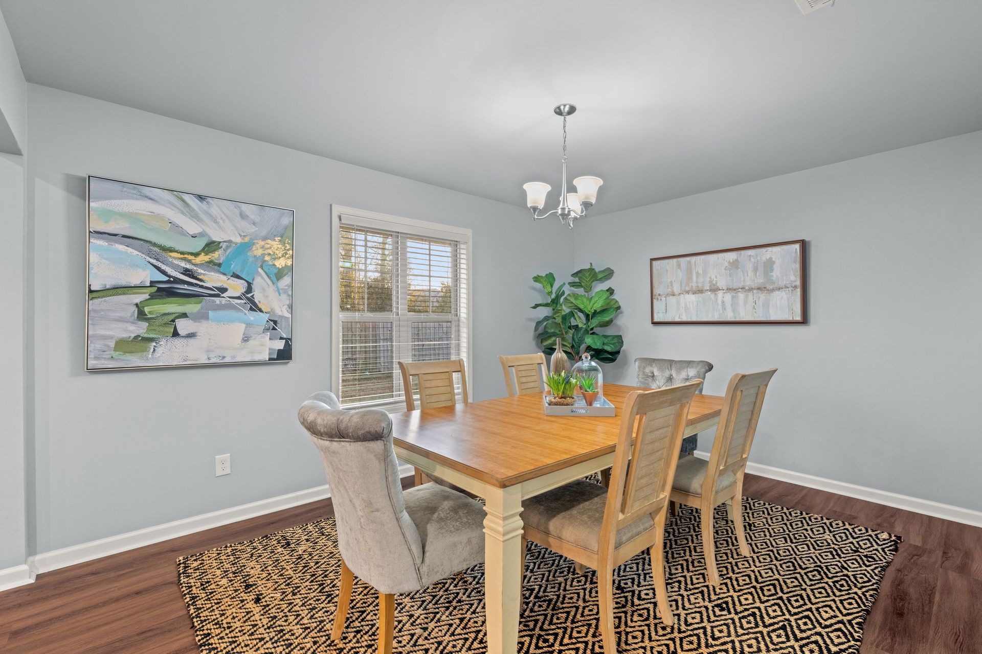 Dining room with wooden table, beige chairs, patterned rug, wall art, and a potted plant.