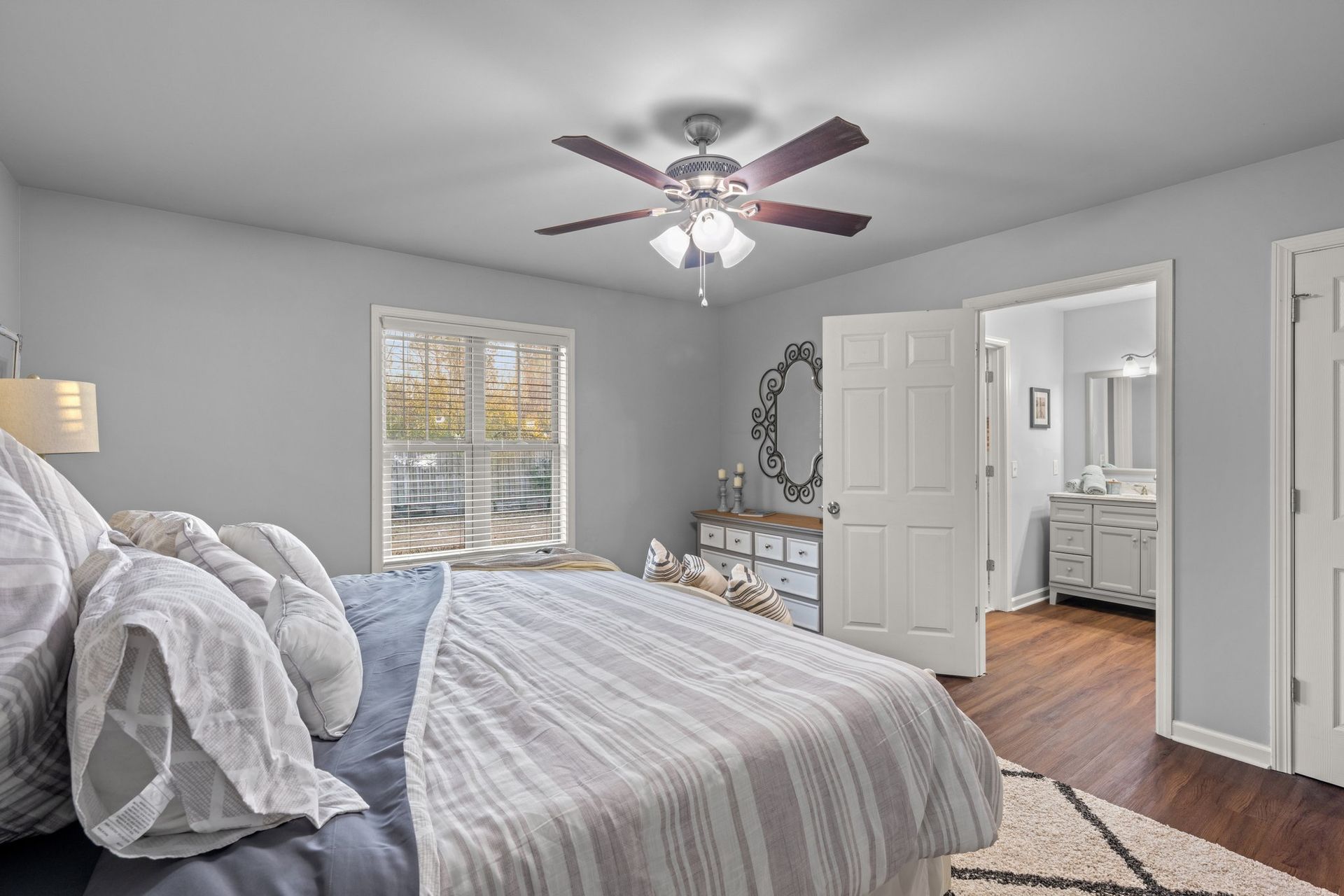 Bright bedroom with gray bedding, ceiling fan, window, and open door to a bathroom.