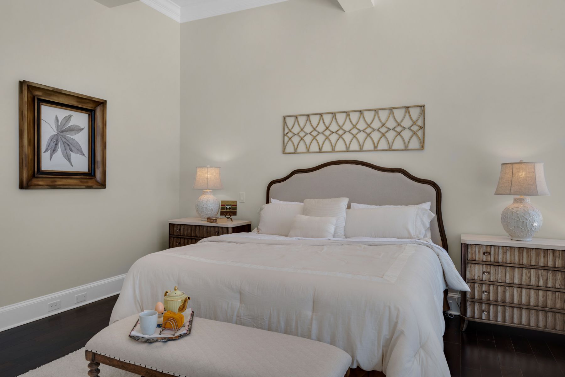 Bright bedroom with a white bed, wicker nightstands, lamps, and beige walls with framed art