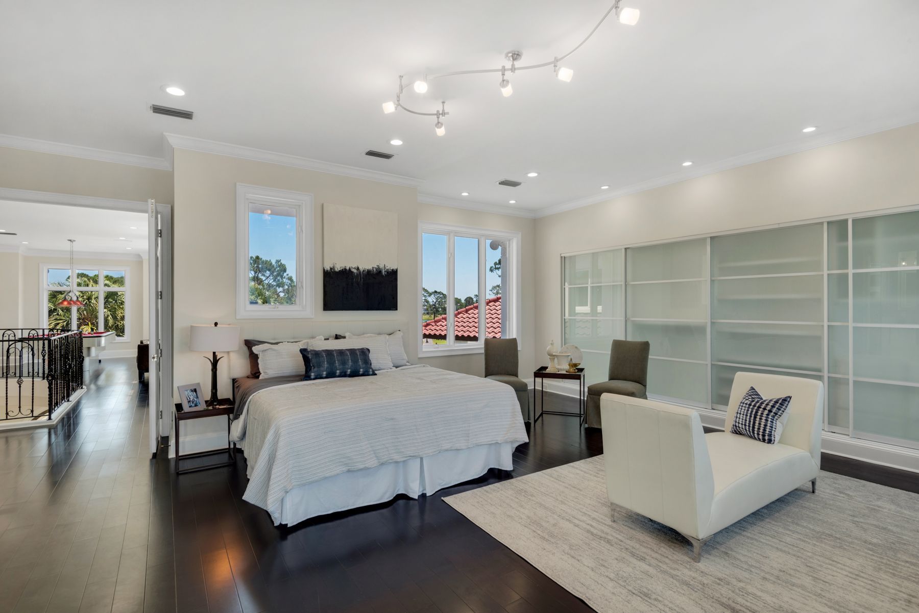 Bright modern bedroom with a white bed, two chairs, large windows, and glass-front shelving