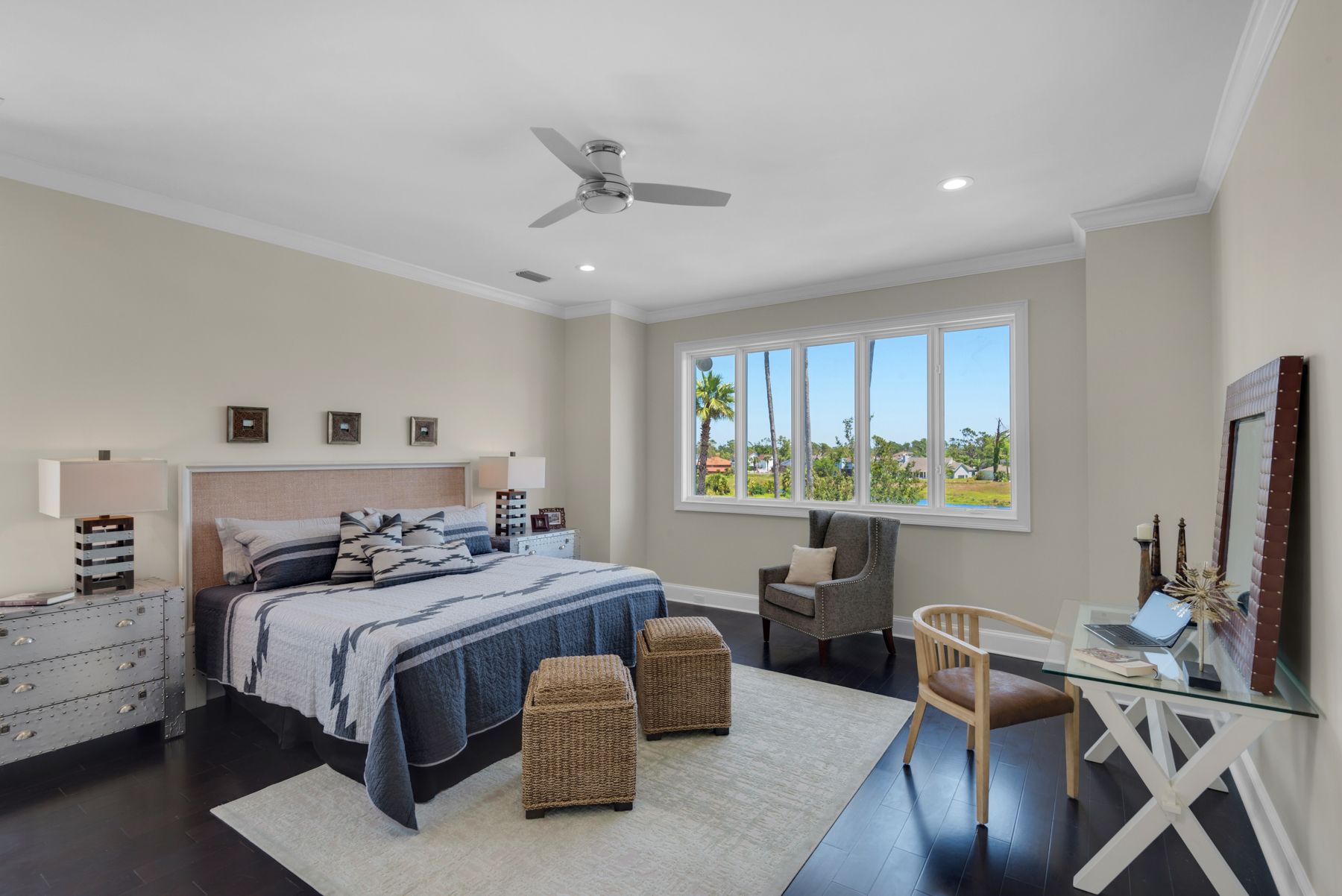 Spacious beige bedroom with a bed, two ottomans, desk, chair, and a large window overlooking greenery