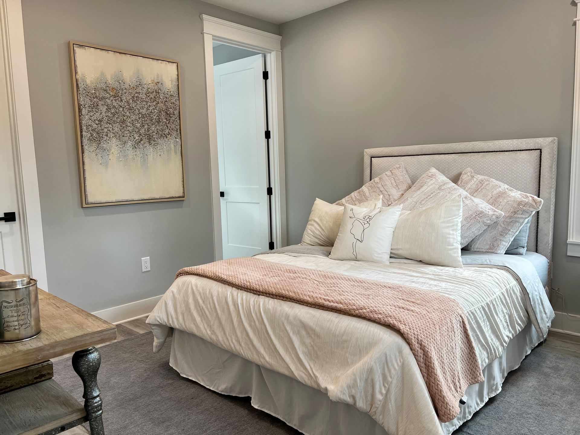 Bright bedroom with a neatly made white bed, soft pink throw, framed wall art, and neutral decor.