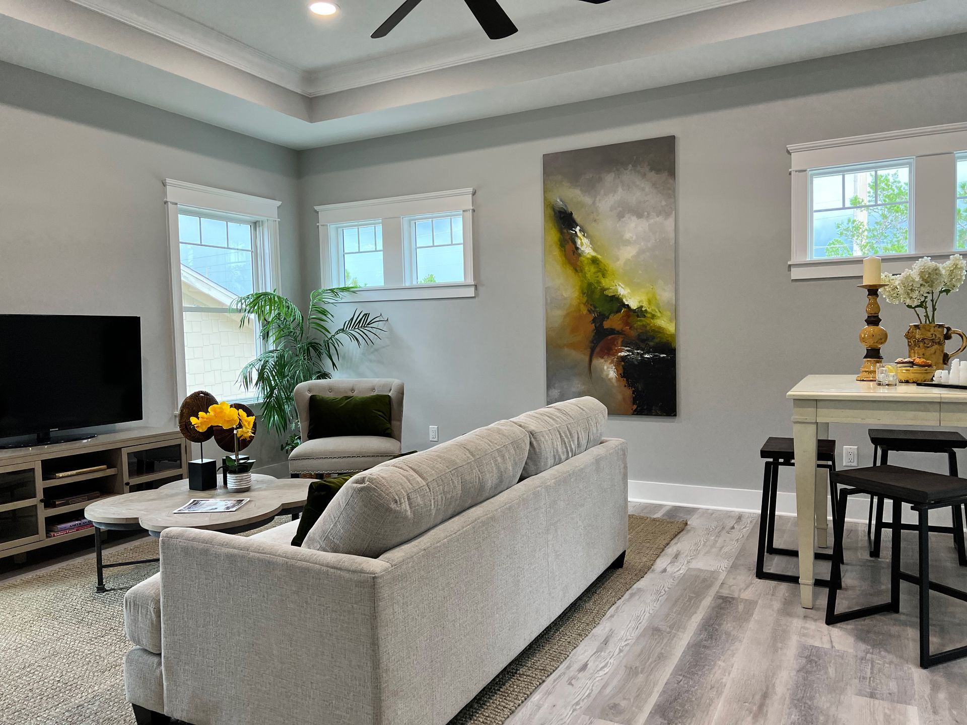 Modern living room with gray sofa, TV, artwork, and dining area by bright windows