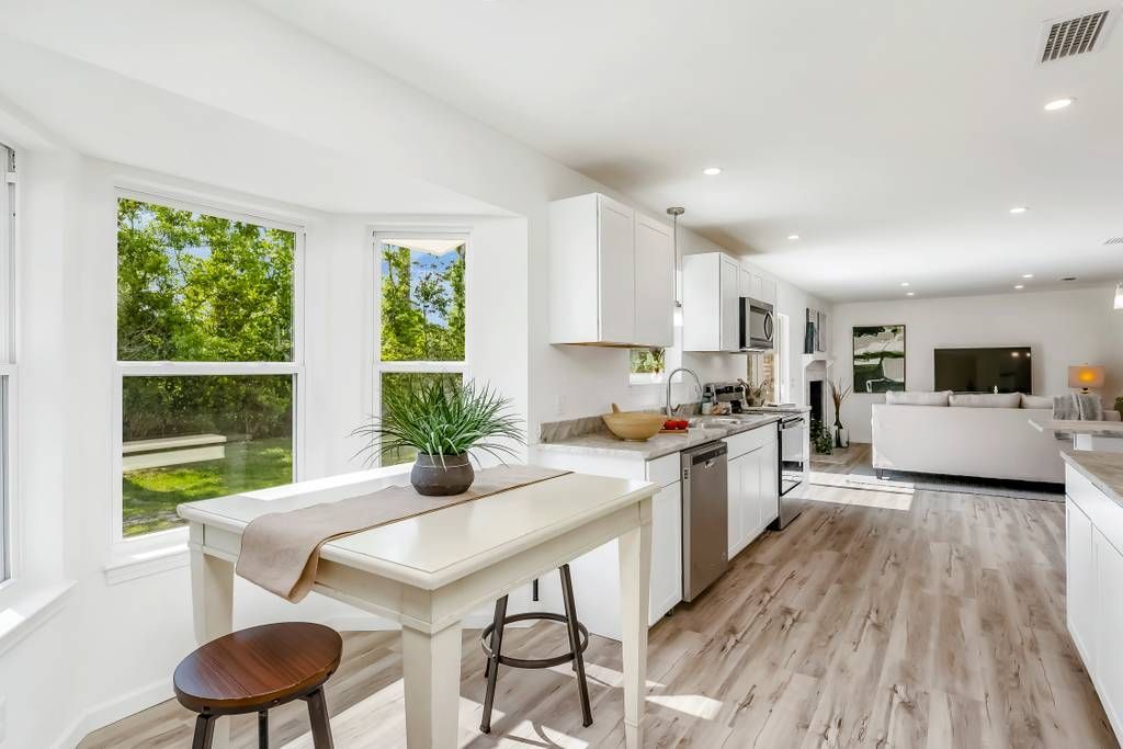 Bright modern kitchen and dining area with white cabinets, wood floors, and large windows overlooking greenery