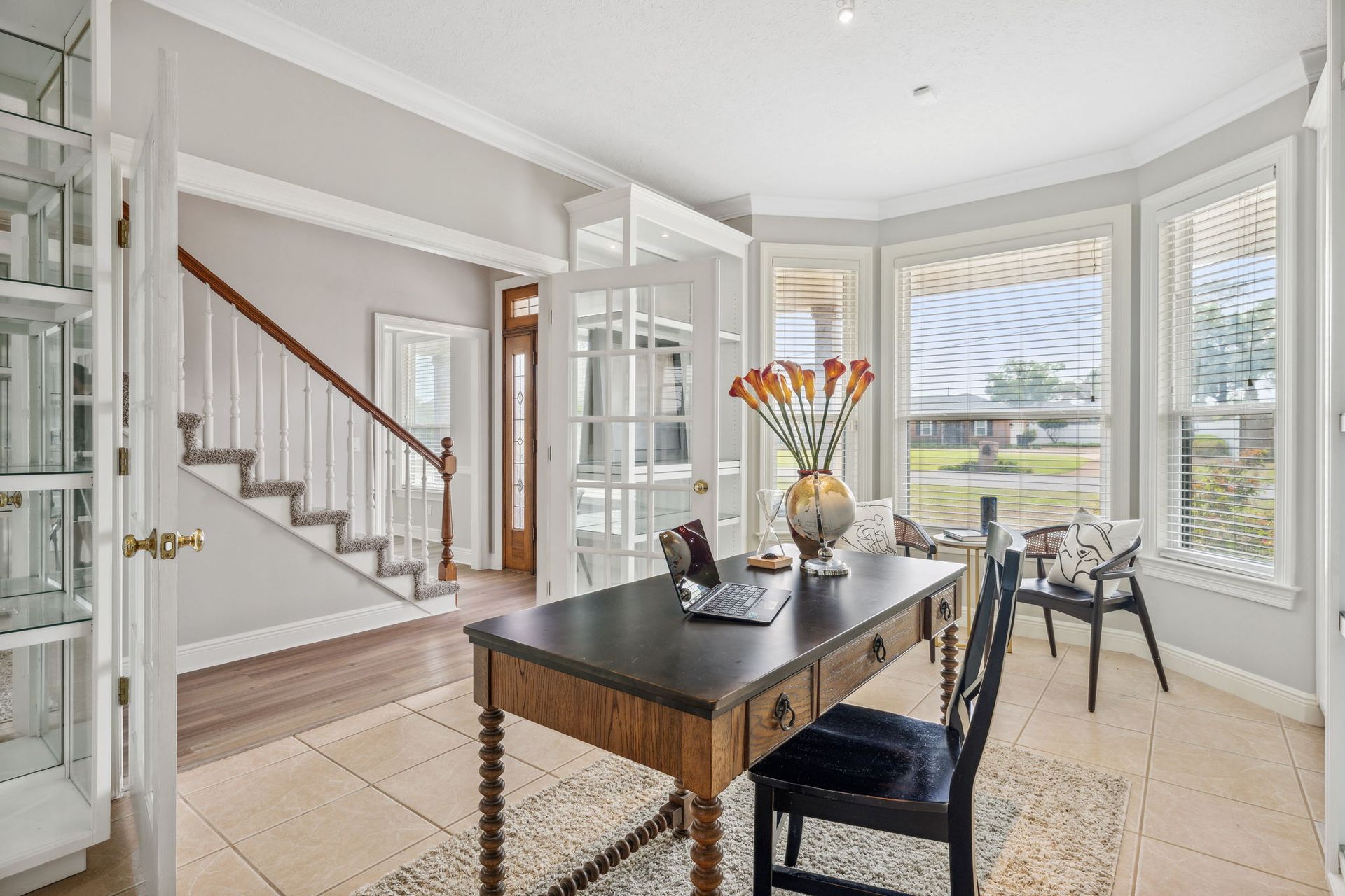 Bright dining room with a dark table, bay windows, staircase, and vase of flowers.