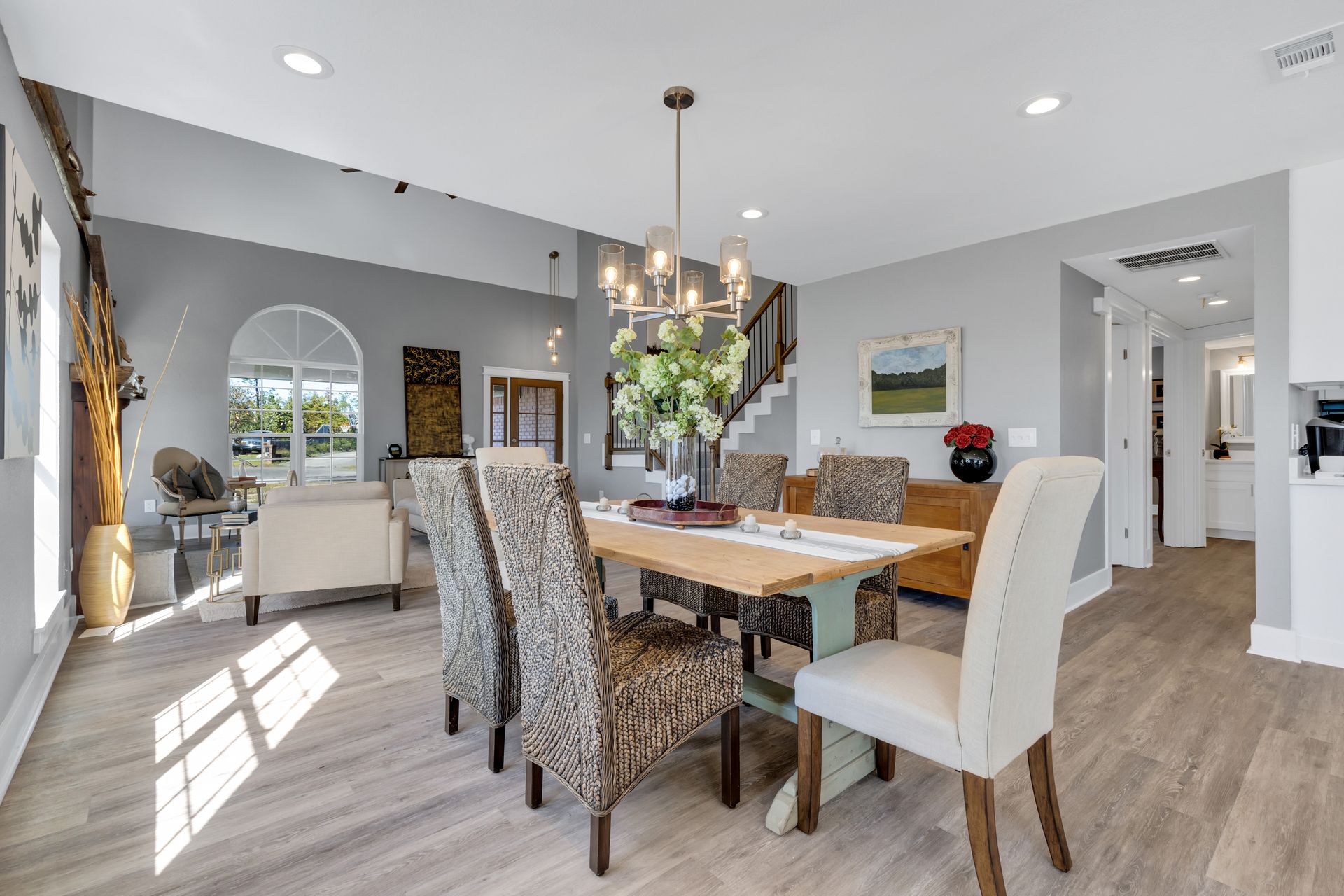 Bright dining room with a long wood table, patterned chairs, chandelier, and sunlit gray walls