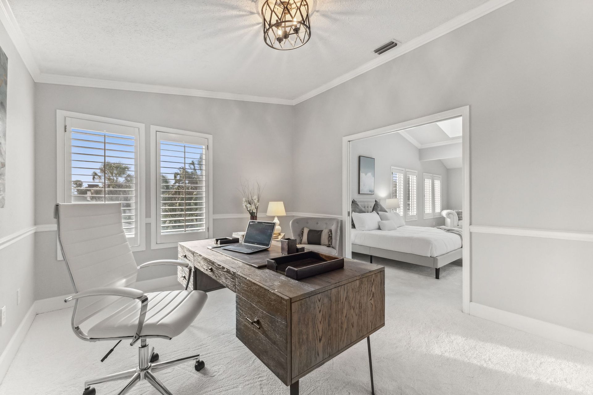 Bright home office with a desk, white chair, windows, and a bedroom visible through an open doorway