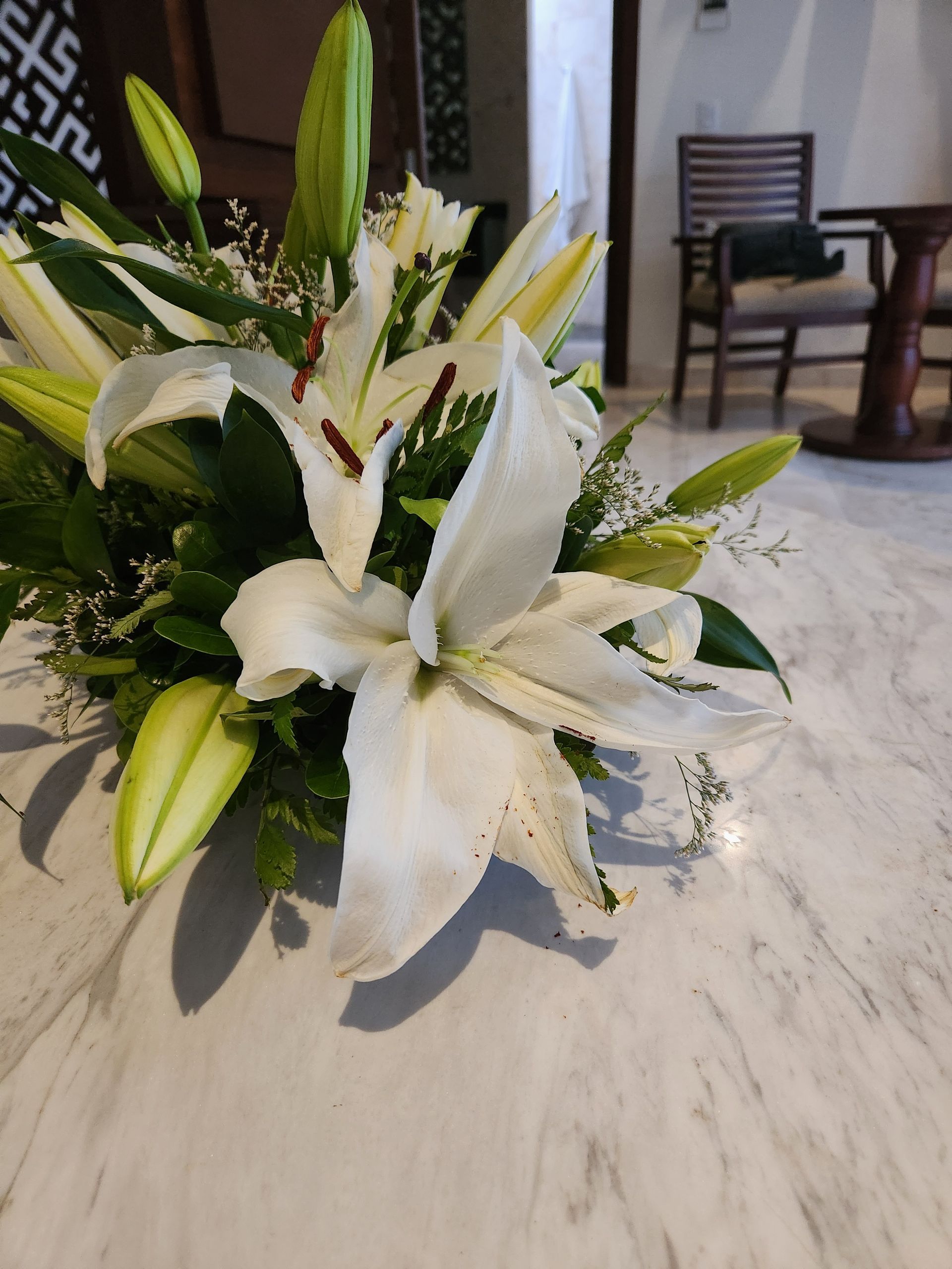 A bouquet of white lilies with green buds and filler flowers sits on a white marble table in a blurred room.