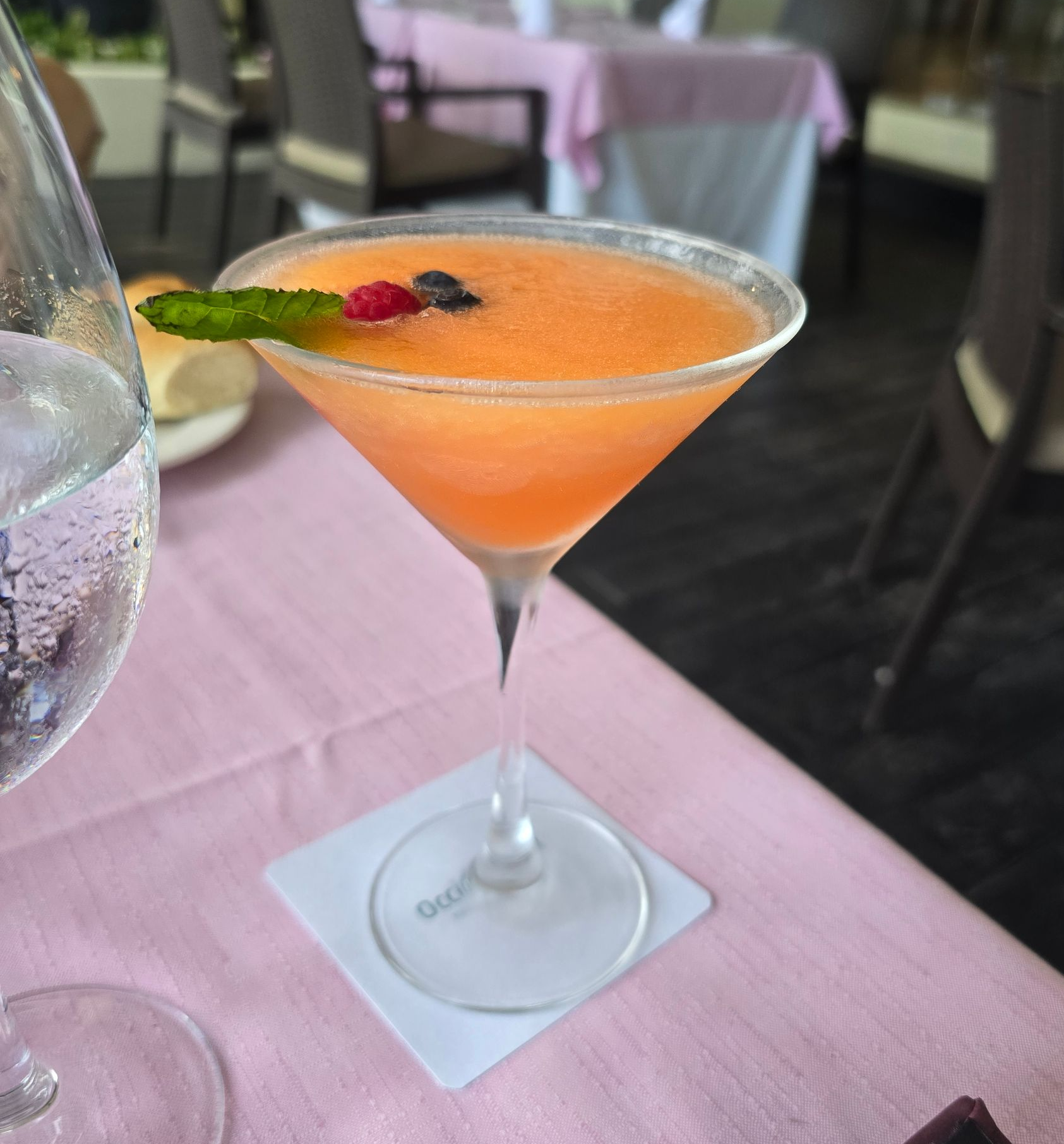 A peach-colored blended cocktail in a martini glass, garnished with a mint leaf and berries, set on a pink tablecloth.