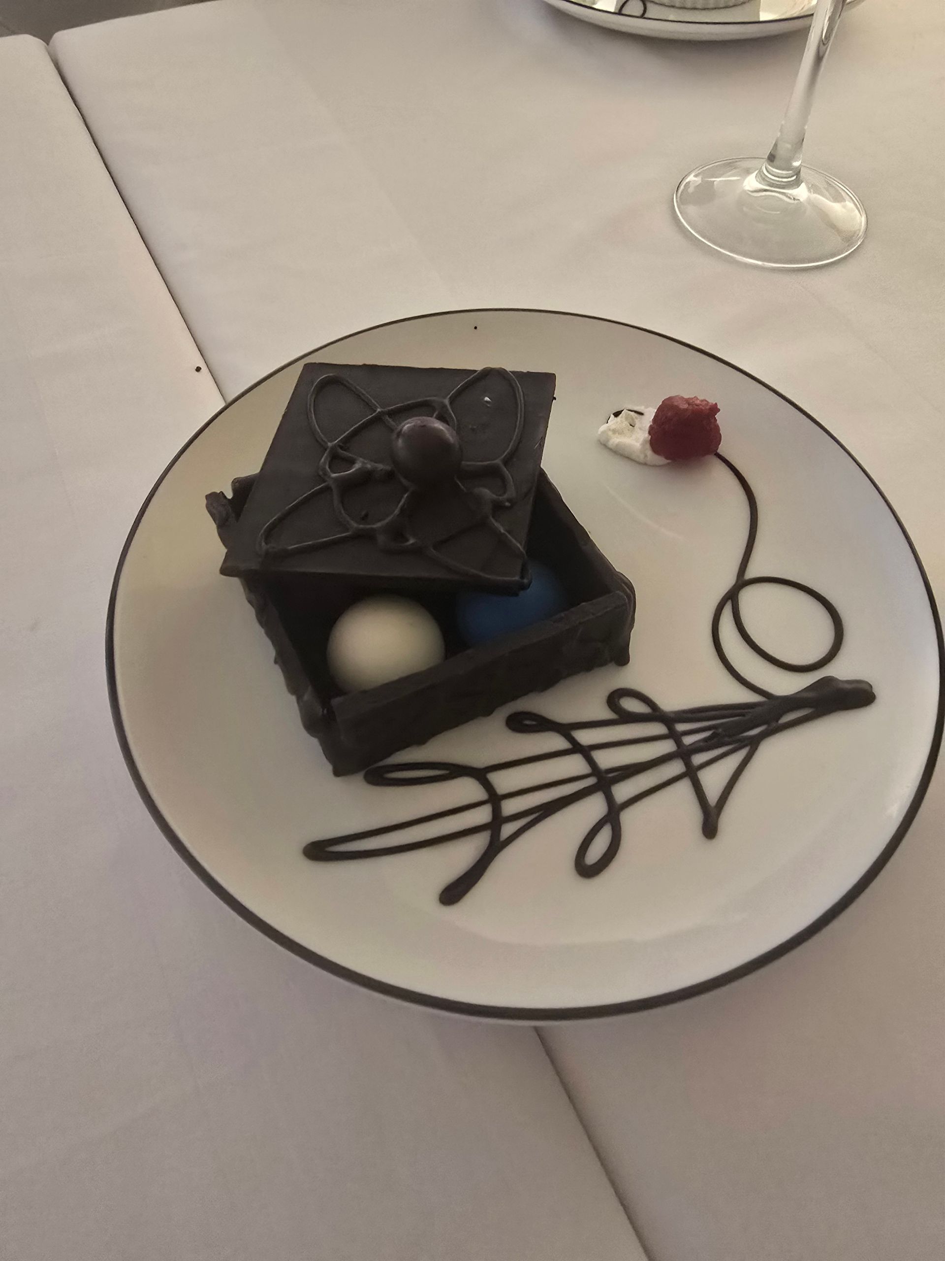 A chocolate box dessert with white and blue truffles inside, served on a white plate with chocolate sauce art and a berry.