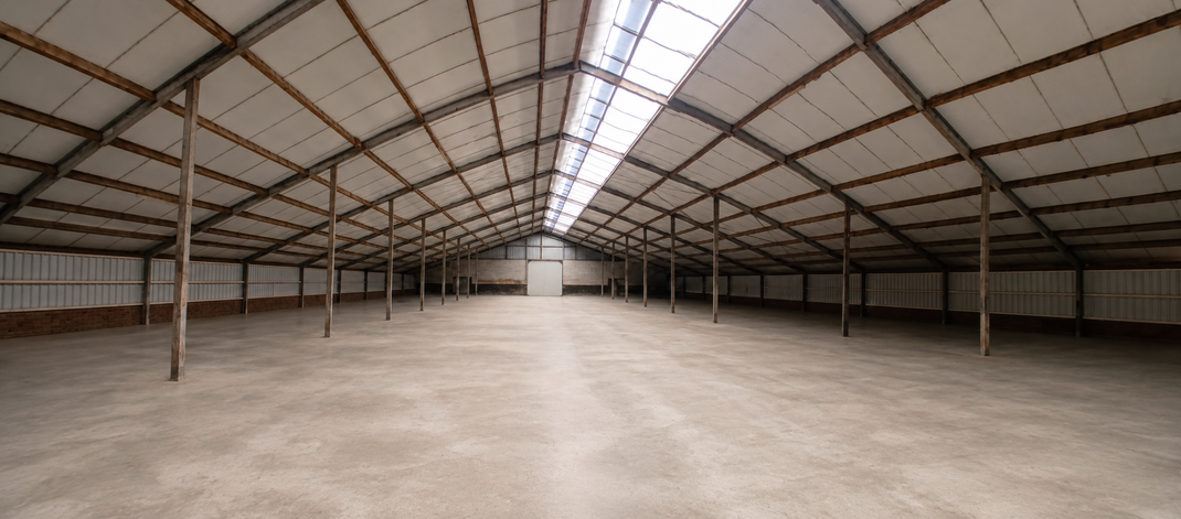 Leeg magazijninterieur met metalen spanten en een brede betonnen vloer.