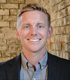 A man in a suit and shirt is smiling in front of a brick wall.