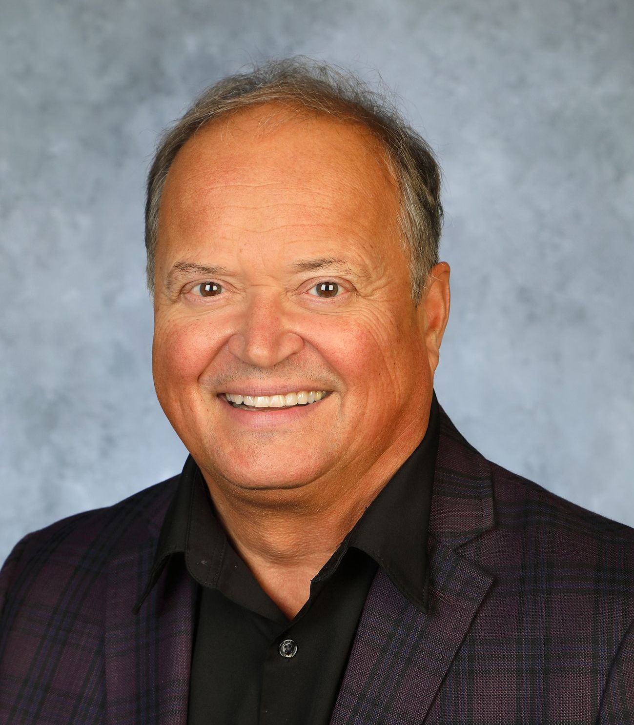 A man in a suit and tie is smiling in front of a purple background.