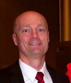 A man in a suit and shirt is smiling in front of a brick wall.