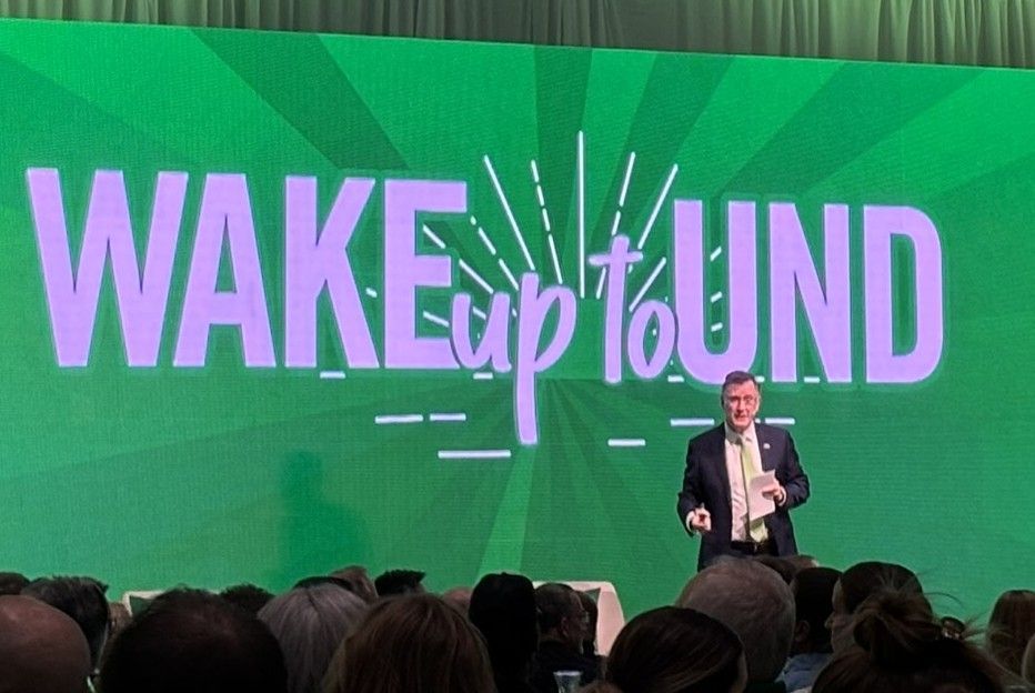 Speaker on stage before green “WAKE up to UND” conference backdrop, audience visible in foreground