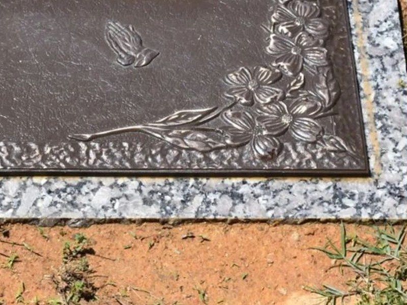Corner of a bronze grave marker with cast dogwood blossoms and praying hands, on a gray granite base
