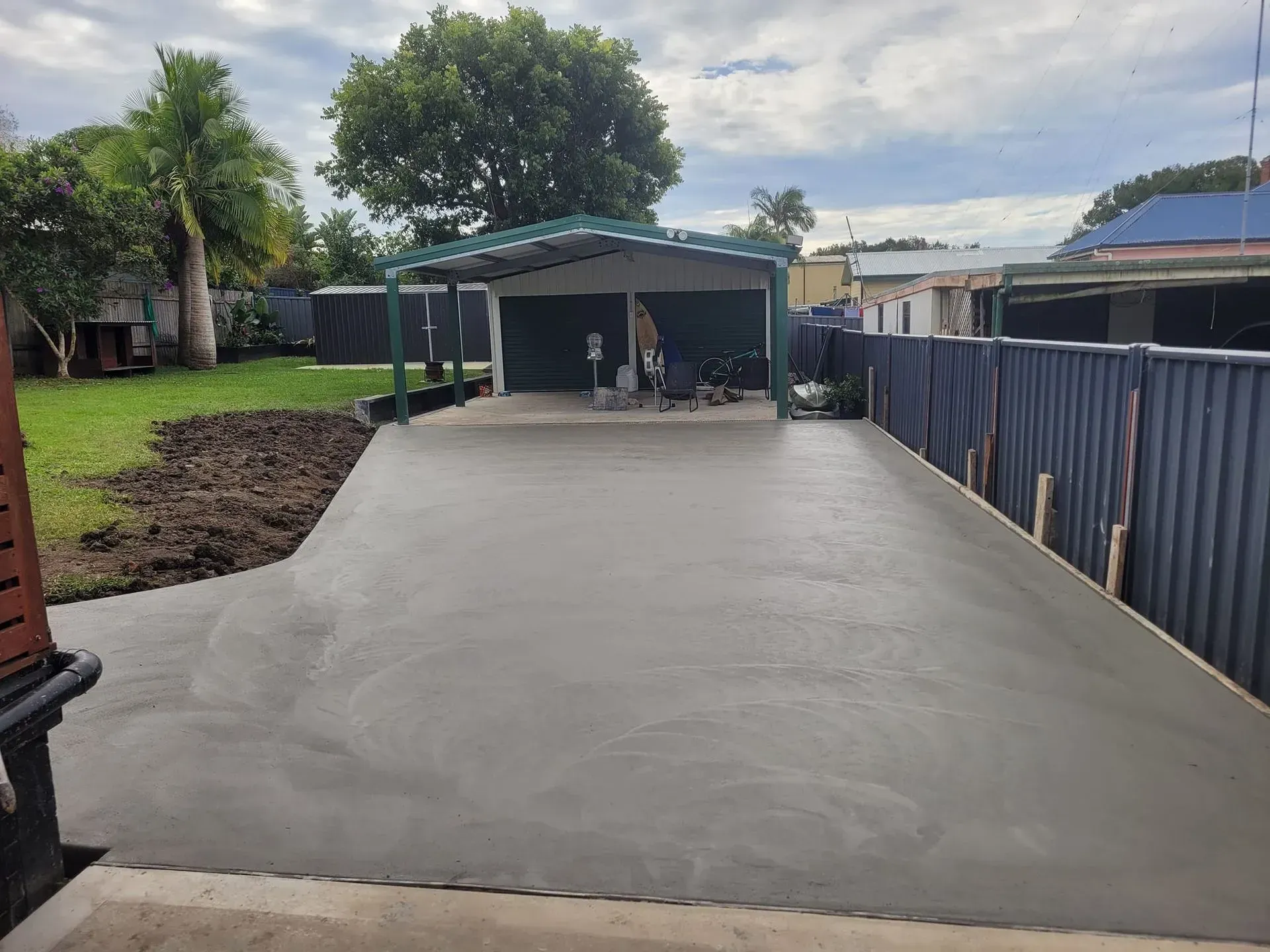Freshly Poured Concrete Driveway Leading to A Carport Beside a Blue Fence and Grassy Yard — Kempsey Concrete Contractors PTY LTD In Dondingalong, NSW