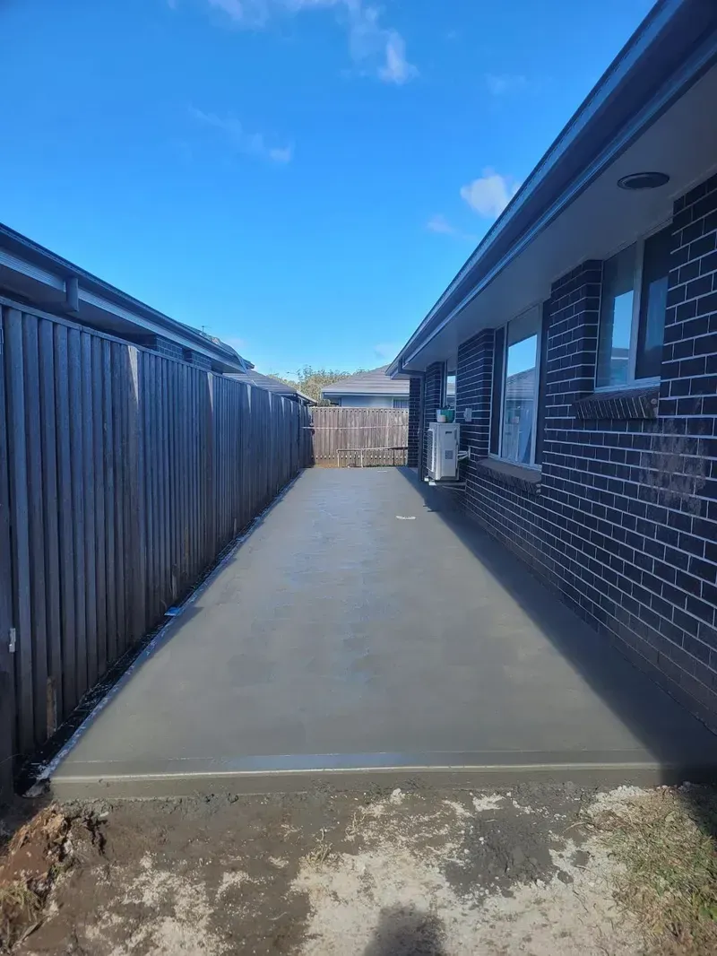 Narrow Concrete Walkway Between a Wooden Fence and Brick House Under a Bright Blue Sky — Kempsey Concrete Contractors PTY LTD In Dondingalong, NSW