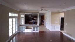 Empty living room with hardwood floors, white trim, large windows, and an open kitchen bar area