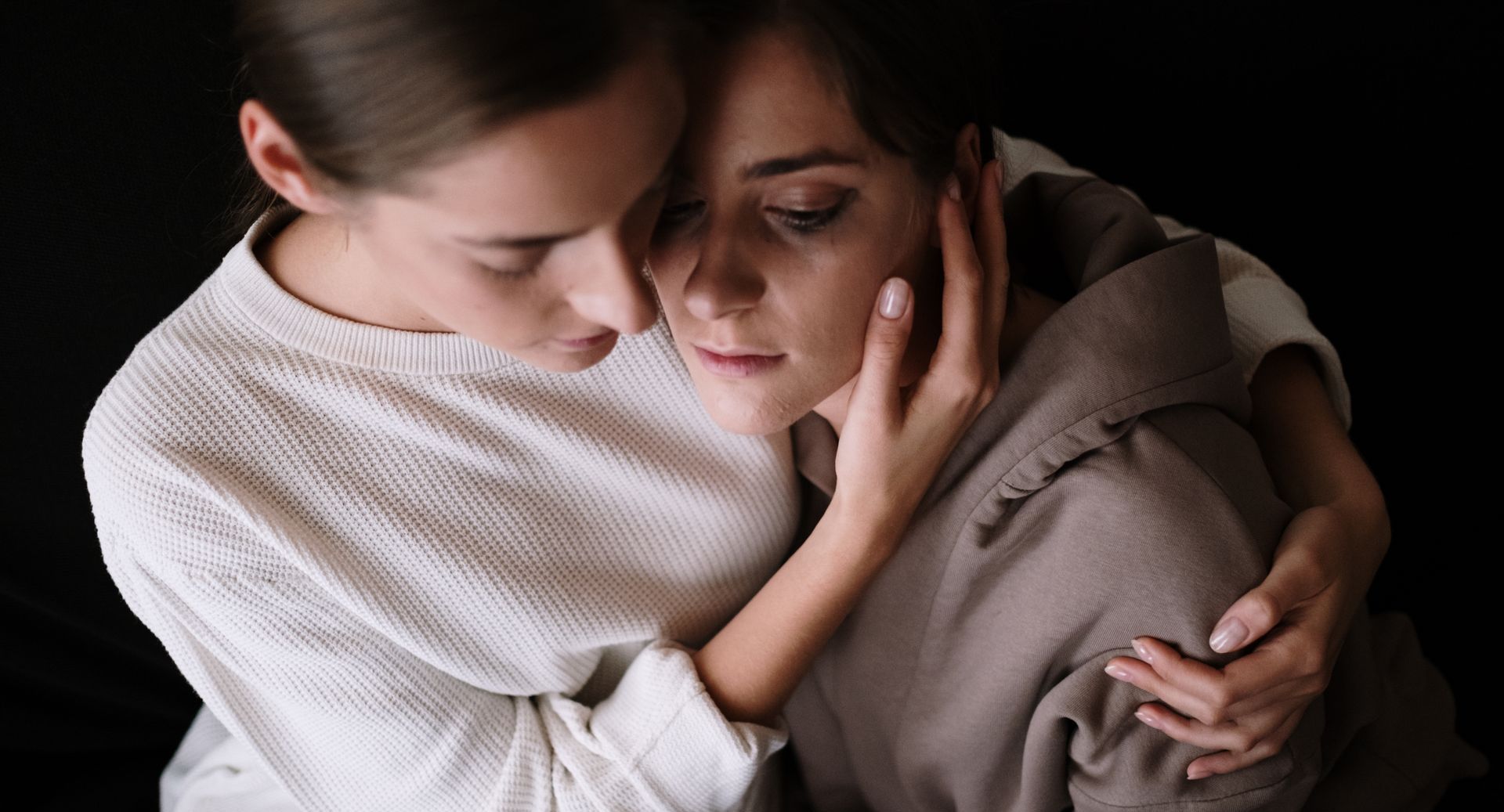 Woman comforts another woman; one's face is in the other's hands, expressing sadness. Both wear sweaters, dark background.