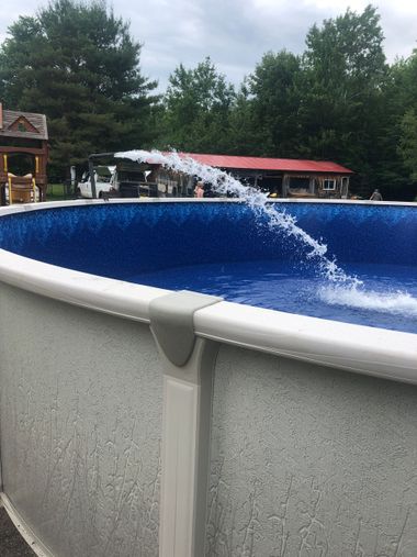 A stream of water sprays from a nozzle into an above-ground swimming pool with a blue liner and gray exterior.