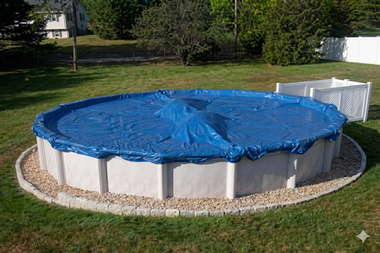A round above-ground pool covered with a blue tarp, surrounded by a gravel border and green lawn.