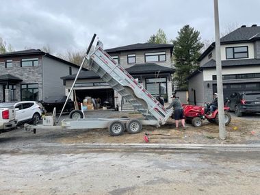 A dump trailer raised in a driveway between suburban homes, with a person operating a red lawn tractor nearby.