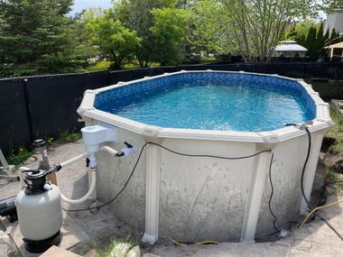 An above-ground oval swimming pool with a filter system, set on a gravel patio against a fence and green trees.