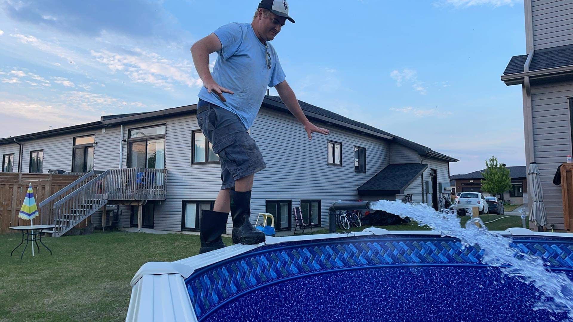 A person in a light blue shirt and dark shorts balances on the edge of a backyard pool while water sprays into the pool.