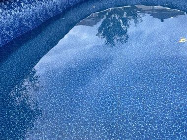 The surface of a swimming pool with a blue patterned liner and reflections of trees and the sky on the water.