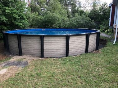 Above-ground circular swimming pool with light tan paneled siding and black vertical supports in a grassy backyard.